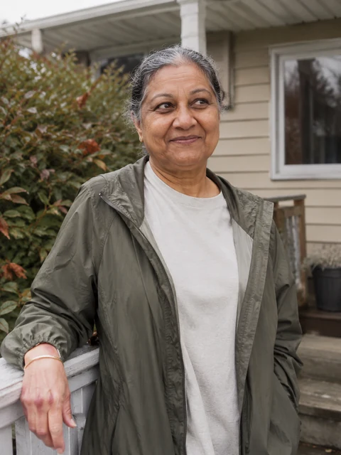 An older woman in an olive jacket resting by her porch railing